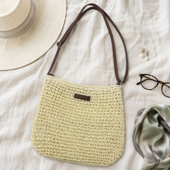 Bolso trenzado en tono crudo con correa marron ajustable, junto a un sombrero de paja y gafas