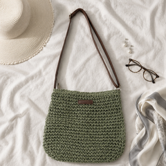 Bolso trenzado en verde salvia con correa marron ajustable, sobre una tela blanca