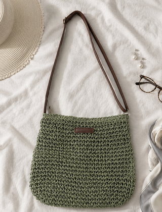 Bolso trenzado en verde salvia con correa marron ajustable, sobre una tela blanca