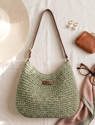 Bolso trenzado en verde salvia sobre lino claro junto a un sombrero, un monedero y gafas