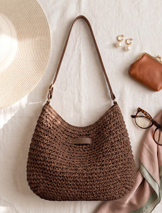 Bolso trenzado en marrón chocolate sobre lino claro junto a un sombrero, un monedero y gafas